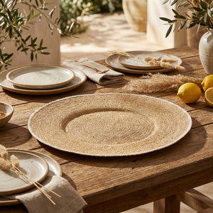 Woven placemat on rustic wooden table with lemons, ceramic plates, and napkins in a sunny setting.
