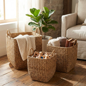 Three woven baskets with blankets, toys, and books on a wooden floor beside a green plant and sofa.