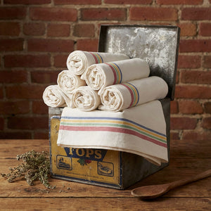 Rolled white towels with colorful stripes in a vintage metal box, on a wooden table. Brick wall backdrop.