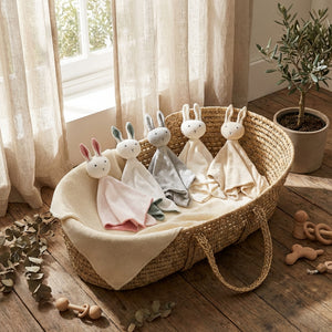 Four plush bunnies in pastel colors sit in a woven basket on a wooden floor near a potted plant.