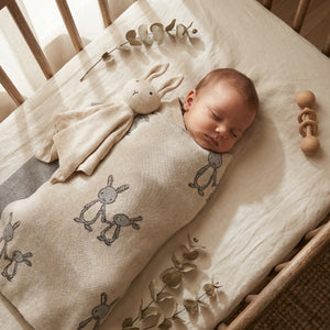 Sleeping baby in a crib, wrapped in a beige blanket with bunny designs, next to a wooden rattle.