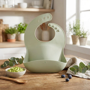 Light green silicone bib with adjustable buttons on a wooden table, near a bowl of green puree.