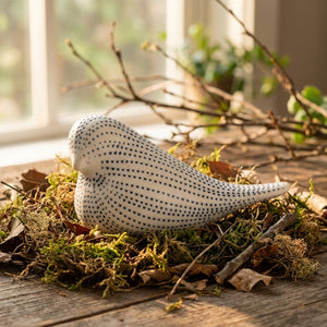 Porcelain bird sculpture with blue dots, resting on moss and leaves on a wooden surface by a window.