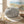 Round, striped vase on sand with driftwood, pebbles, and wheat stalks, against a blurred beach scene.