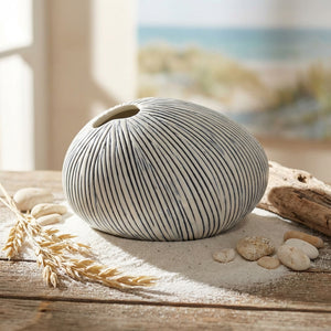 Round, striped vase on sand with driftwood, pebbles, and wheat stalks, against a blurred beach scene.