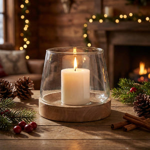 White candle in glass holder on wooden table, surrounded by pinecones and greenery, cozy background.
