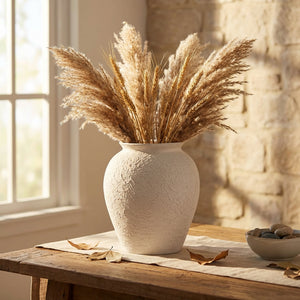 White vase with pampas grass on wooden table, sunlit stone wall, window light streaming in.