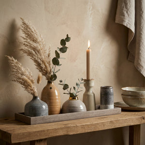 Vases with pampas and eucalyptus on a wooden tray beside a lit candle and ceramic cups on a bench.