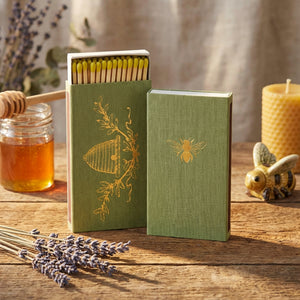 Green matchbox with gold bee design, open to reveal yellow-tipped matches, lavender, honey jar nearby.