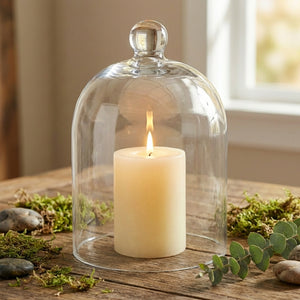 White candle under a glass dome on a wooden table, surrounded by green moss and smooth stones.