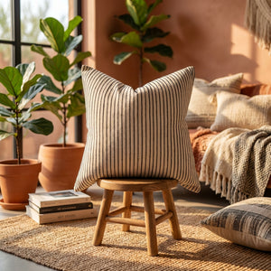 Striped pillow on wooden stool, surrounded by potted plants and books on a woven rug.