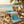 Beach scene with a wooden table, blue towel, straw bag, sunscreen, hat, sunglasses, and palm trees.