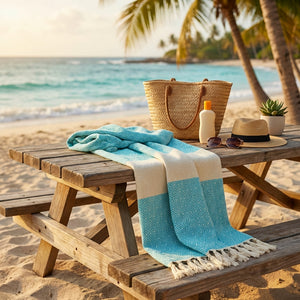 Beach scene with a wooden table, blue towel, straw bag, sunscreen, hat, sunglasses, and palm trees.