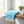 Blue towel with white stripes and tassels on a wooden shelf, next to a plant and glass jar.