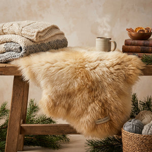 Cozy scene with a fluffy beige throw, knit blankets, steaming mug, books, and yarn on a wooden table.