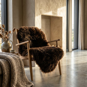 Cozy chair with brown fur in sunlit room, dried flowers in a vase, and knitted blanket on a sofa.