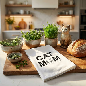 Cat figurine, fresh herbs, rustic bread, and "Cat Mom" towel on a wooden board in a cozy kitchen.
