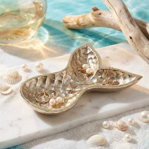 Gold mermaid tail dish with jewelry, pearls, and seashells on a sandy, marble surface by water.