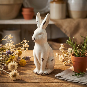 White ceramic rabbit with distressed finish on wooden table, surrounded by dried flowers and a plant.