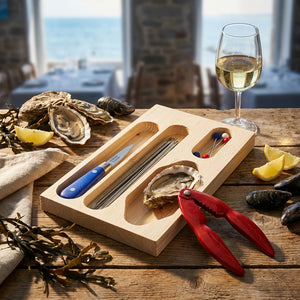 Oyster shucking kit with knife, forks, and red tool on wood table, sea view, wine glass nearby.