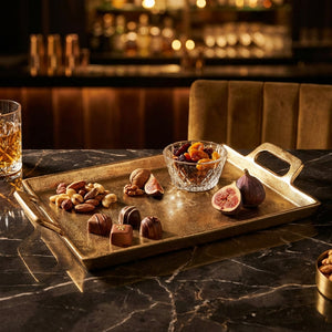 Golden tray with nuts, chocolates, figs, and a glass bowl of dried fruits on a dark marble table.