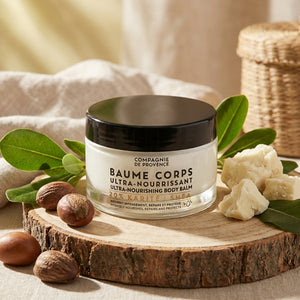 Body balm jar on wood slab, surrounded by green leaves, nuts, and shea butter in soft lighting.