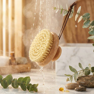 Wooden brush with bristles, water dripping, eucalyptus leaves, and stones on a marble surface.