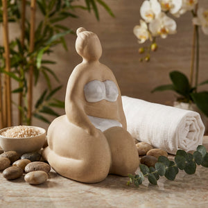 Beige sculpture of a woman with white swimsuit, surrounded by stones, towel, and orchids.
