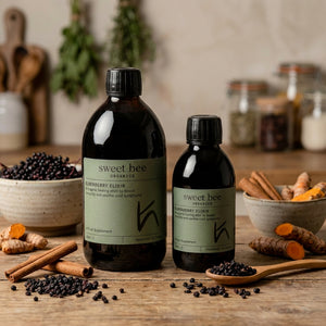 Two dark bottles labeled "Sweet Bee" on a wooden table with elderberries, turmeric, and cinnamon.