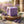 Purple soap cube on wooden slab, surrounded by lavender and olive leaves, in sunlit field.