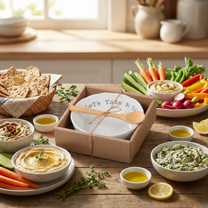 A wooden table with colorful vegetables, dips, a bowl set, crackers, and small bowls of olive oil.