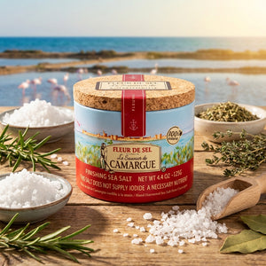 Tin of Fleur de Sel on a wooden table with salt, rosemary, and herbs, sea and flamingos in background.