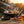 Barbecue grill with meat, vegetables, and tools at sunset; beer and herbs on wooden table.