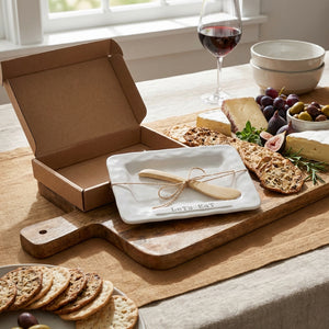 Cheese platter with figs, crackers, and olives on a wooden board; wine glass and open box nearby.