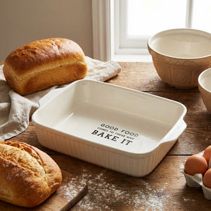 Bread, bowls, and eggs on a wooden table with a white baking dish reading "Good food...BAKE IT."