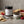 Jar of raspberry pepper preserve with crackers, bowl of spread, raspberries, and peppercorns.