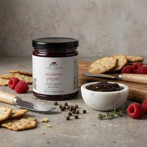Jar of raspberry pepper preserve with crackers, bowl of spread, raspberries, and peppercorns.