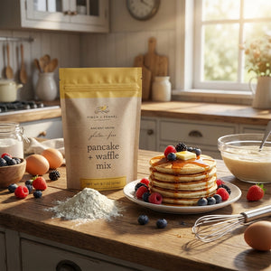 Pancakes with berries and syrup on a table, alongside pancake mix, flour, eggs, and a whisk.