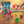 Hand tossing a red ball among colorful jacks on wood floor, with jumbo jax set packaging in view.