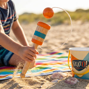 Child playing with a wooden toy on a sandy beach, colorful striped blanket, and a beach bucket nearby.