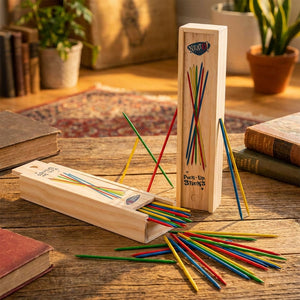 Colorful pick-up sticks scattered from a wooden box on a rustic table with books and a plant.