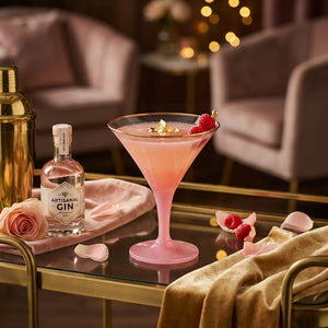 Pink cocktail in a glass with raspberries and gold flakes, on a table with gin bottle and rose petals.