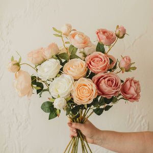A hand holds a bouquet of pink, white, and peach roses against a textured, off-white background.