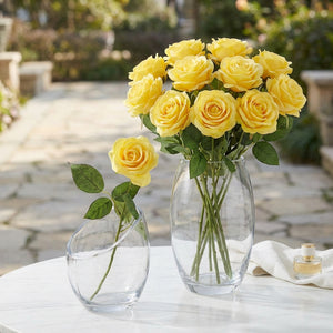 Yellow roses in clear vases on a white table, with a blurred stone path and greenery in the background.