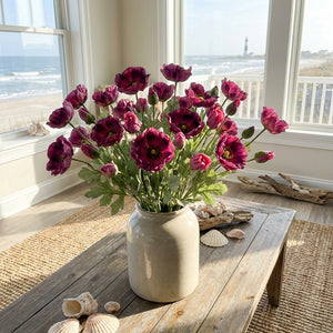 Vibrant pink flowers in a vase on a wooden table by the beach, lighthouse visible through windows.