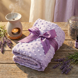 Lilac textured blanket with purple ribbon, lavender sprigs, candle burner, and essential oil on wood.