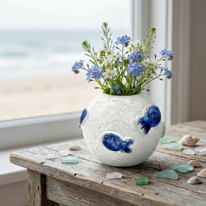 White vase with blue fish design, holding blue and white flowers, on a wooden table by a window.