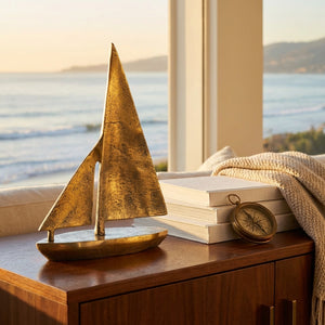 Golden sailboat sculpture on a wooden table by a window, with books and a compass, ocean view.