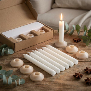 White candles with holders on a wooden table, surrounded by green leaves and star anise.