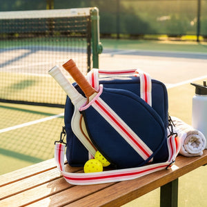 Blue bag with white and red straps, pickleball paddles, yellow balls on bench by tennis court.